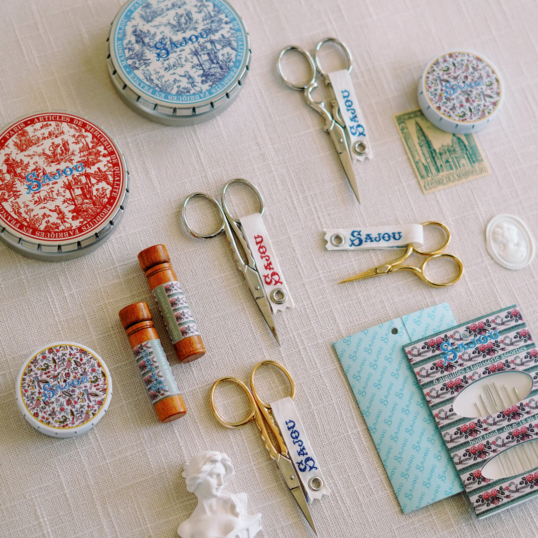 Flat lay of Maison Sajou needlepoint accessories styled on linen, featuring scissors, tins, and vintage needle booklets.