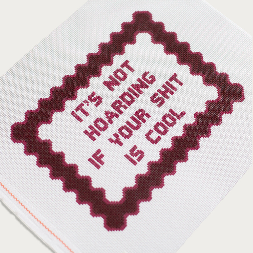 needlepoint canvas with maroon text in all caps that reads "It's not hoarding if your shit is cool" and a dark maroon frame