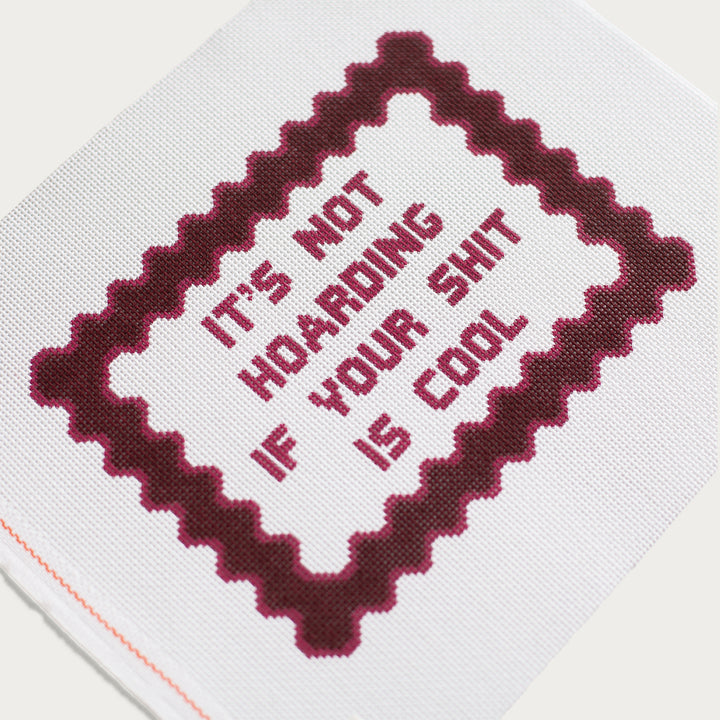 needlepoint canvas with maroon text in all caps that reads "It's not hoarding if your shit is cool" and a dark maroon frame