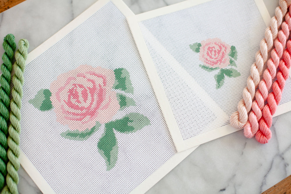 two needlepoint canvases beside one another. one with a larger pink rose and the other with a smaller one. pink and green threads frame the edges