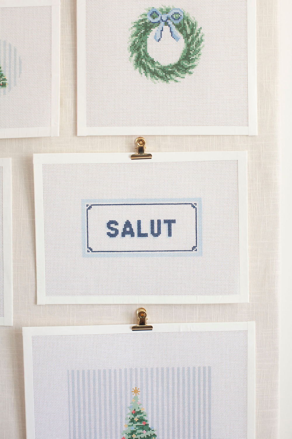 a light cream bulletin board with needlepoint canvases clip to it with gold clips. the center canvas says salut in blue and the one above it is a green wreath with a blue bow