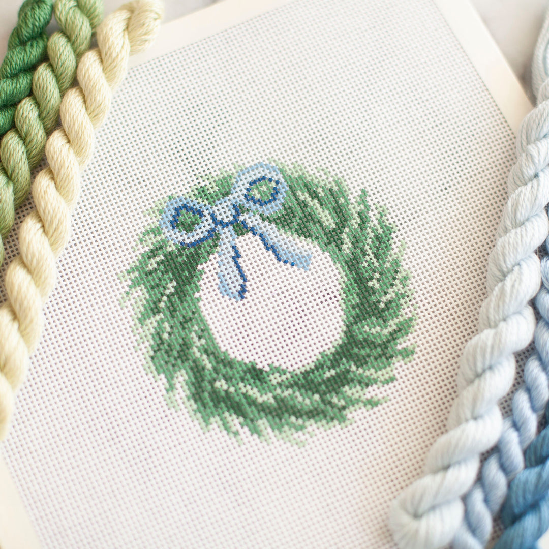 Needlepoint Canvas with a Christmas Wreath with Blue Bow hanging from the top. green and blue skiens of thread are on either side of the canvas